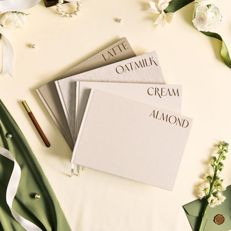 K&ouml;nnte beinhalten: Vier Notizb&uuml;cher mit den W&ouml;rtern "LATTE", "OATMILK", "CREAM" und "ALMOND" auf den Einb&auml;nden. Die Notizb&uuml;cher sind in Beige- und Cremet&ouml;nen gehalten. Ein goldfarbener Stift und eine Perlenkette sind ebenfalls zu sehen.