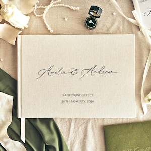 Könnte beinhalten: Ein cremefarbenes Hochzeitsgästebuch mit den Namen "Amelia & Andrew" in eleganter Schrift. Das Buch ist von Hochzeitsaccessoires umgeben, darunter eine Ringbox, Perlen und Blumen. Der Text "SANTORINI GREECE 26TH JANUARY, 2026" ist darunter gedruckt.