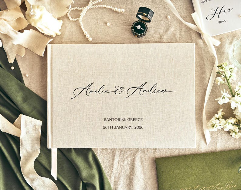 K&ouml;nnte beinhalten: Ein cremefarbenes Hochzeitsg&auml;stebuch mit den Namen "Amelia & Andrew" in eleganter Schrift. Das Buch ist von Hochzeitsaccessoires umgeben, darunter eine Ringbox, Perlen und Blumen. Der Text "SANTORINI GREECE 26TH JANUARY, 2026" ist darunter gedruckt.