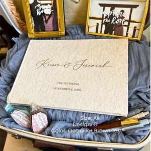 K&ouml;nnte beinhalten: Eine Hochzeits-Themenausstellung mit einem Fotoalbum, zwei gerahmten Fotos und zwei Rollen Klebeband. Das Album ist cremefarben mit braunem Text "Krissa & Jeremiah" und "The Pittmans, November 2, 2025."