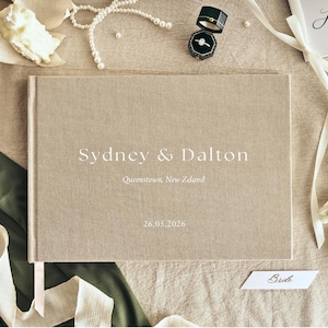 K&ouml;nnte beinhalten: Ein beiges Hochzeitsg&auml;stebuch aus Leinen mit den Namen "Sydney & Dalton" und dem Datum "26.05.2026" auf dem Einband. Das Buch ist von Hochzeitsaccessoires umgeben, darunter Ringe, Blumen und B&auml;nder.