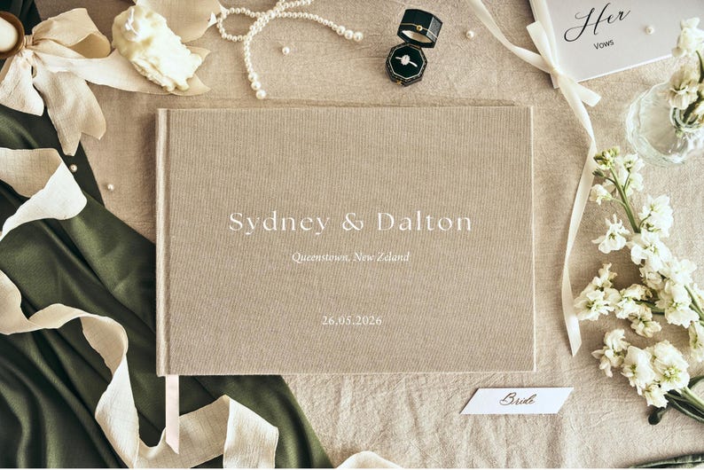 K&ouml;nnte beinhalten: Ein beiges Hochzeitsg&auml;stebuch aus Leinen mit den Namen "Sydney & Dalton" und dem Datum "26.05.2026" auf dem Einband. Das Buch ist von Hochzeitsaccessoires umgeben, darunter Ringe, Blumen und B&auml;nder.