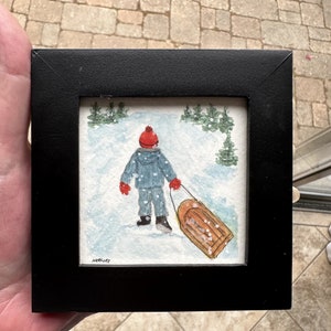 May include: A watercolor painting of a child in a red hat and blue coat pulling a wooden sled through a snowy landscape. The painting is framed in black.