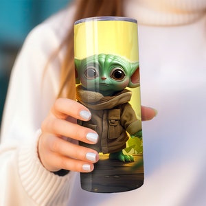 May include: A yellow and green stainless steel tumbler with a cartoon image of a green alien character wearing a brown jacket. The alien is standing on a wooden surface.