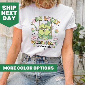 May include: White t-shirt with a green four-leaf clover cartoon character swinging on a swing. The text "One Lucky Occupational Therapist" is printed on the shirt.