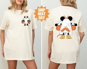 May include: A white t-shirt with a graphic of a ghost wearing a Minnie Mouse costume and holding pumpkins. The ghost is wearing a red bow and has black eyes. The text "Ship Next Day" is visible in the background.