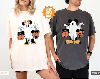 May include: Two t-shirts with a ghost holding jack-o'-lanterns. The ghost is wearing a bow and has Mickey Mouse ears. The text "Ship Next Day" is on a yellow star sticker. The t-shirts are in the colors ivory and pepper.