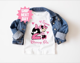 May include: A white t-shirt featuring a Minnie Mouse graphic with the number 2 in pink polka dots. The text "Sarah" and "Birthday Girl" are also in pink. A denim jacket is draped over the shirt.
