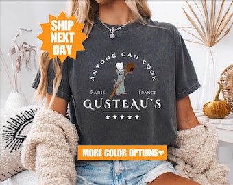 May include: A dark gray t-shirt with a white graphic of a rat wearing a chef's hat and holding a spoon. The text on the shirt reads "Anyone Can Cook" with "Paris" and "France" on either side of the rat. The text "Gusteau's" is below the rat with five stars underneath. The shirt has a distressed look.