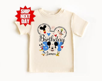 Puede incluir: Una camiseta color crema con un diseño gráfico colorido. El diseño incluye las palabras "Birthday Boy" con los personajes de Mickey Mouse, Pato Donald y Goofy. También se incluye el nombre "James".