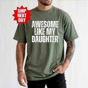 May include: Olive green t-shirt with the text "AWESOME LIKE MY DAUGHTER" in white, distressed font. A red starburst graphic with the words "SHIP NEXT DAY" is in the upper left corner. The shirt is a casual style.
