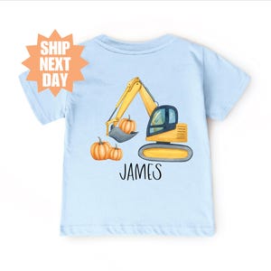 Pode incluir: Camiseta azul clara com uma ilustração de uma escavadeira amarela com abóboras. O nome "JAMES" está impresso em preto. Um design divertido e sazonal para crianças.
