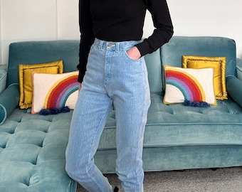 May include: A person wearing a black mock neck shirt and light blue high-waisted jeans with a striped pattern. The person is standing in front of a teal blue sofa with two rainbow patterned cushions.