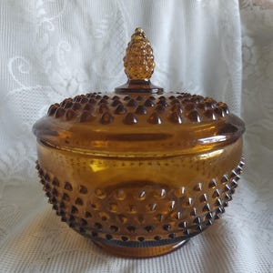 May include: Amber-colored glass candy dish with a lid. The dish has a hobnail pattern and a small, cone-shaped handle on the lid. The dish is round and sits on a small base. The glass has a warm, golden hue.