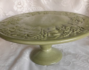 May include: A green ceramic cake stand with a raised design of grapes and leaves. The stand has a round base and a pedestal.