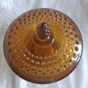 May include: Amber-colored glass jar with a lid. The lid and jar are covered in small, raised, circular bumps. The lid has a small, textured knob in the center. The jar is sitting on a white, lacy surface.