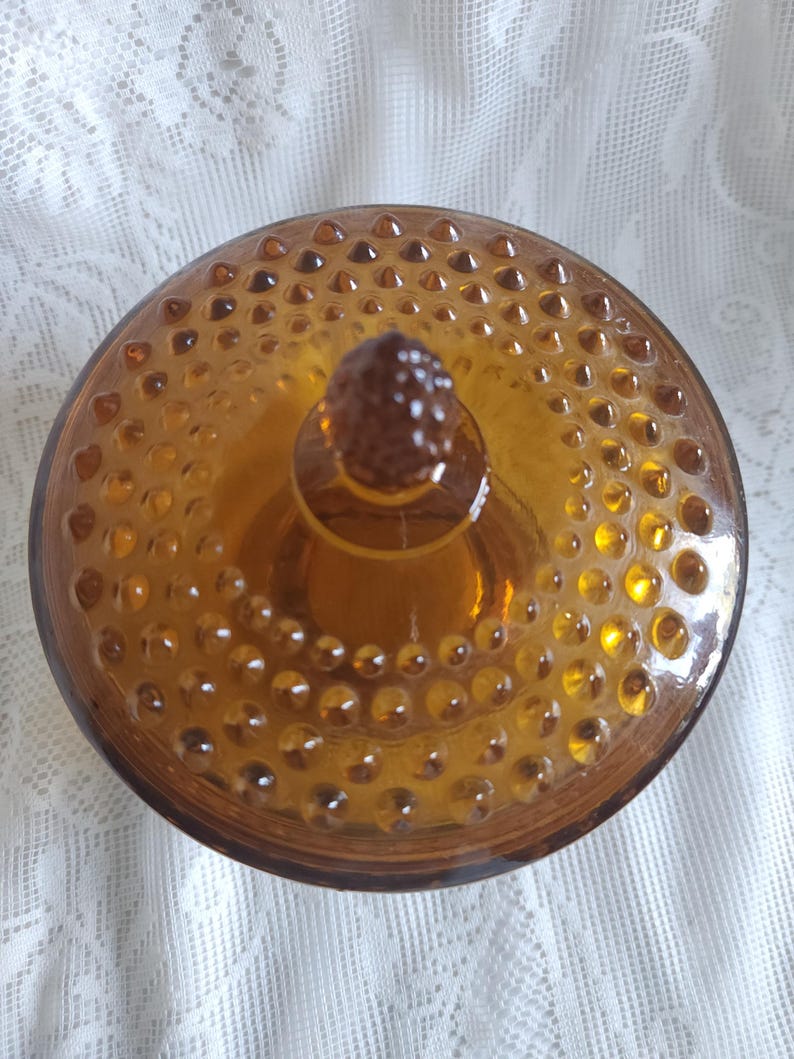 May include: Amber-colored glass jar with a lid. The lid and jar are covered in small, raised, circular bumps. The lid has a small, textured knob in the center. The jar is sitting on a white, lacy surface.