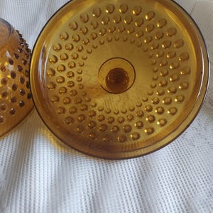 May include: Close-up of a vintage amber-colored glass bowl with a textured, hobnail pattern. The bowl has a round base and a wide, shallow dish shape. The glass has a warm, golden hue.