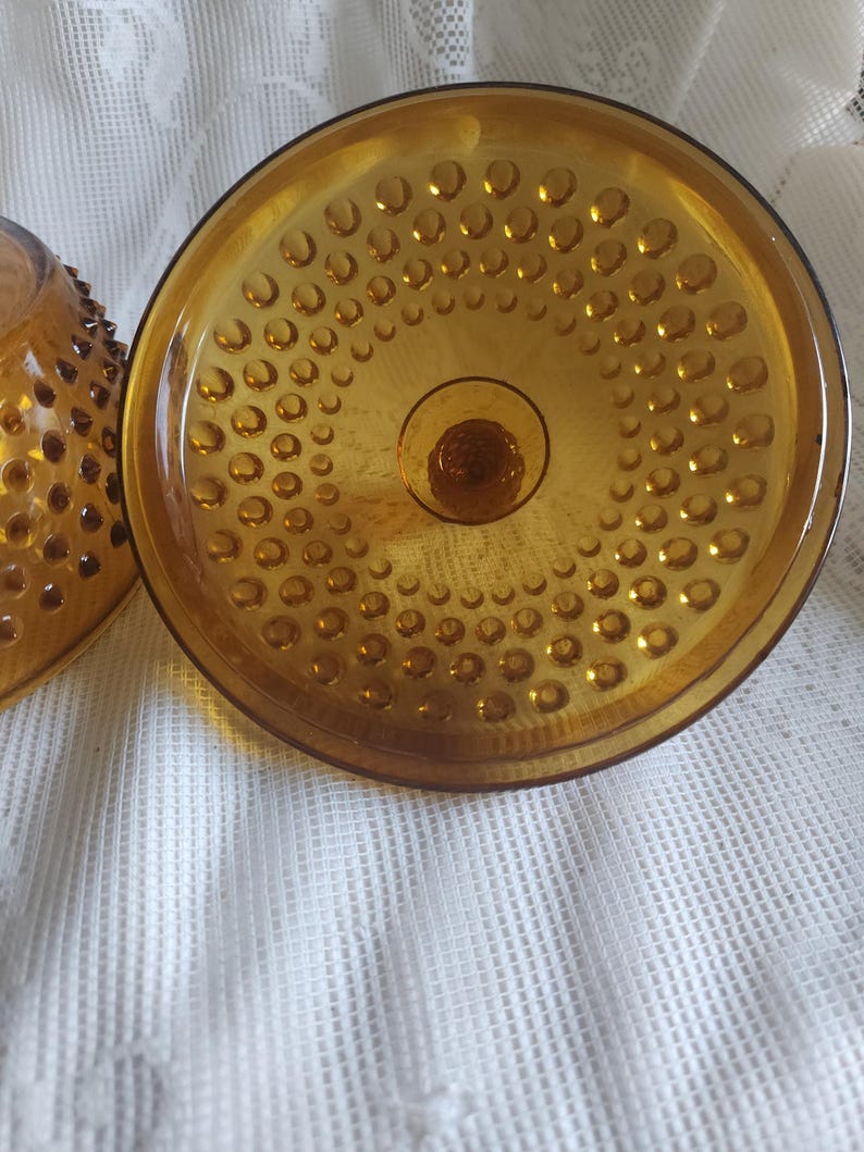 May include: Close-up of a vintage amber-colored glass bowl with a textured, hobnail pattern. The bowl has a round base and a wide, shallow dish shape. The glass has a warm, golden hue.