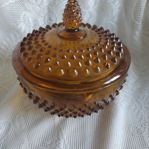 May include: Amber-colored glass candy dish with a lid. The dish has a hobnail pattern and a pinecone-shaped finial on the lid. The dish is round and sits on a white lace surface.