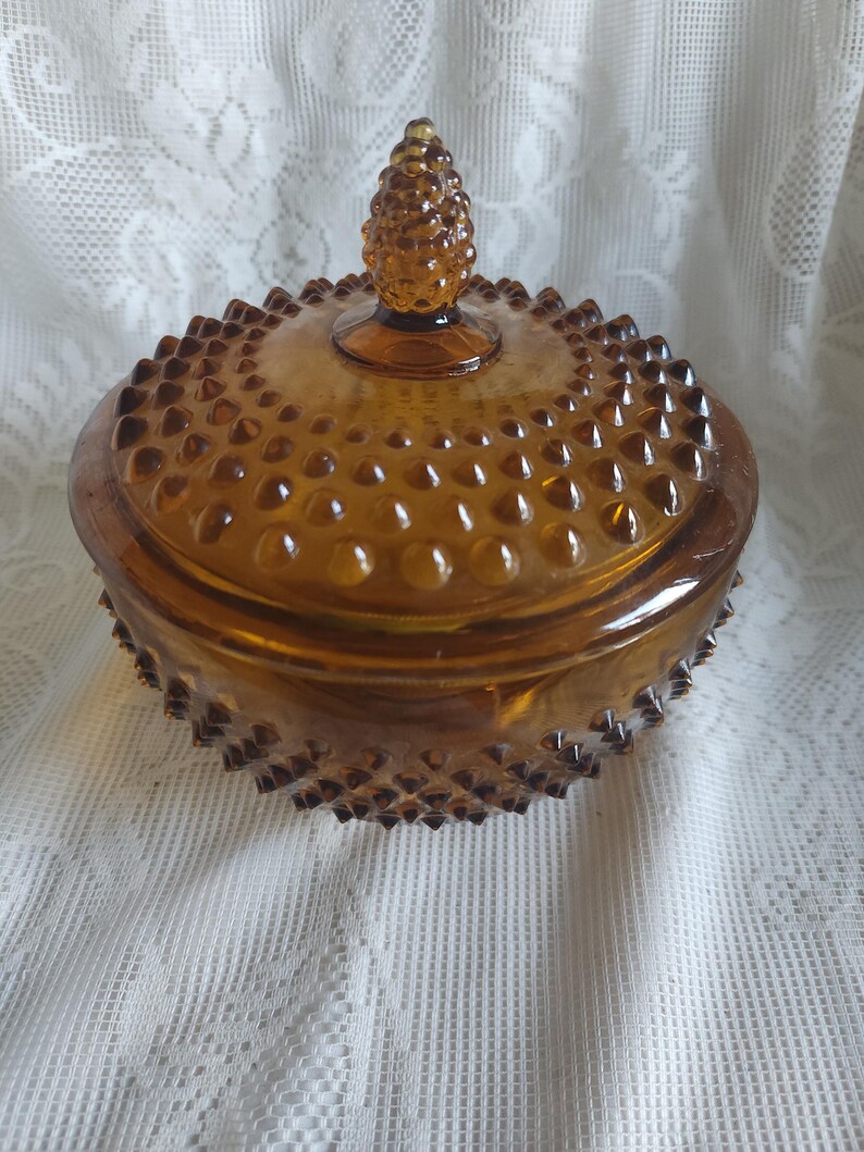 May include: Amber-colored glass candy dish with a lid. The dish has a hobnail pattern and a pinecone-shaped finial on the lid. The dish is round and sits on a white lace surface.