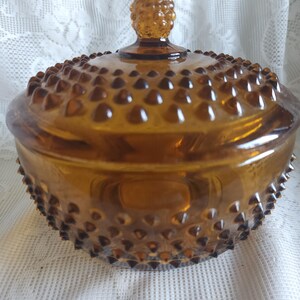 May include: Amber-colored glass candy dish with a lid. The dish has a hobnail pattern and a textured surface. The lid is topped with a decorative finial. The dish is round and sits on a white lace surface.