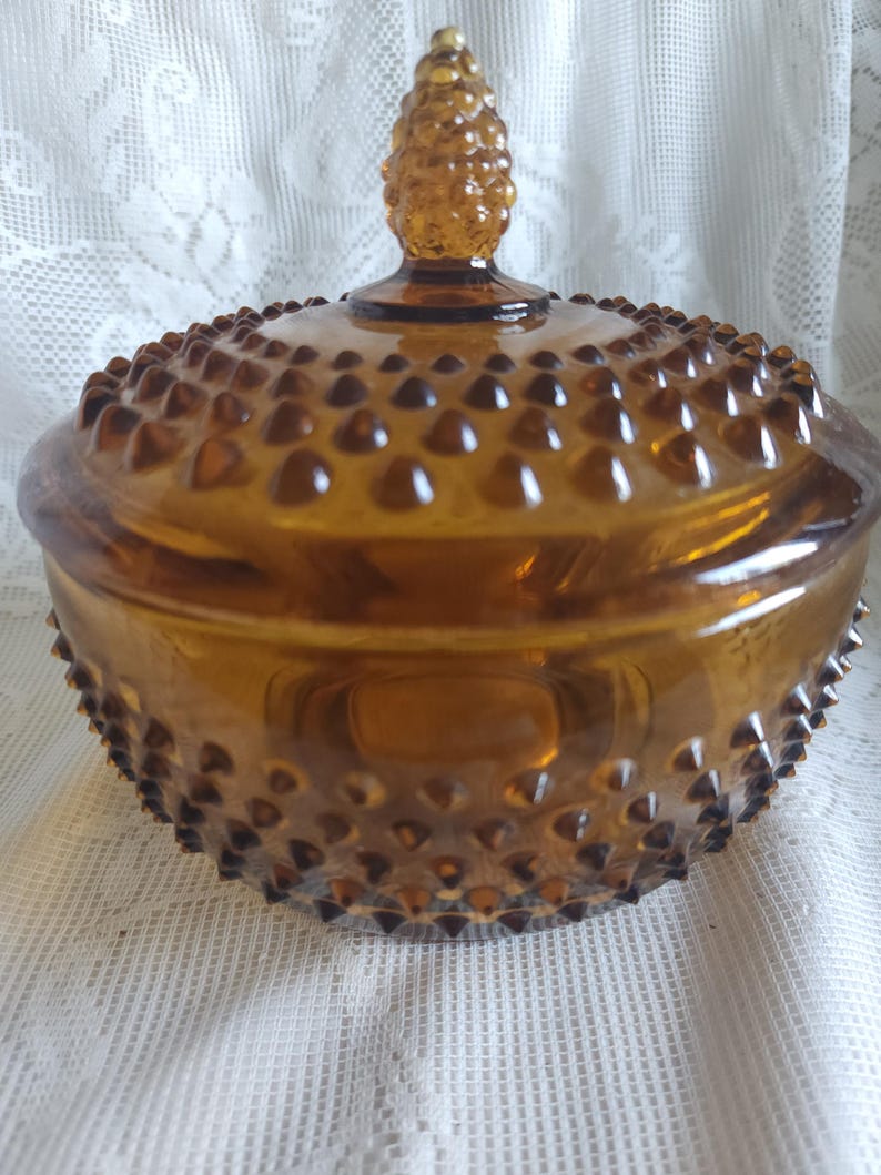 May include: Amber-colored glass candy dish with a lid. The dish has a hobnail pattern and a textured surface. The lid is topped with a decorative finial. The dish is round and sits on a white lace surface.