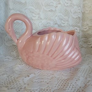 May include: A pink ceramic swan planter with a curved neck and a ruffled edge. The planter has a textured, feathered design on the body. The swan is a light pink color and is likely used for decorative purposes.
