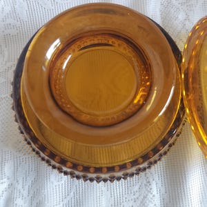 May include: Amber-colored glass dish with a lid. The dish has a round shape with a textured pattern on the base and a decorative rim. The lid is also amber-colored and has a similar design. The dish is sitting on a white, textured surface.