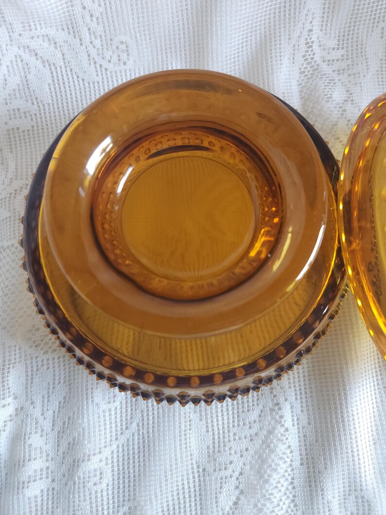 May include: Amber-colored glass dish with a lid. The dish has a round shape with a textured pattern on the base and a decorative rim. The lid is also amber-colored and has a similar design. The dish is sitting on a white, textured surface.