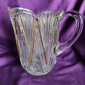 May include: A clear glass pitcher featuring a scalloped rim and a vertical fan-like pattern with gold-toned accents. The body of the pitcher displays a textured design with alternating smooth and beaded vertical lines, complemented by a curved handle.