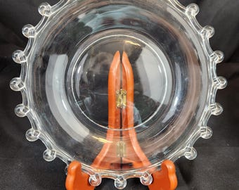 May include: A clear glass plate with a decorative beaded edge, displayed on a wooden stand. The plate is round and has a slightly raised center. The wooden stand is a warm orange color and has a folding design.