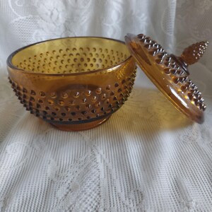 May include: Amber-colored glass candy dish with a hobnail pattern. The lid is off, revealing the interior. The dish sits on a white lace tablecloth. The lid has a knob on top.