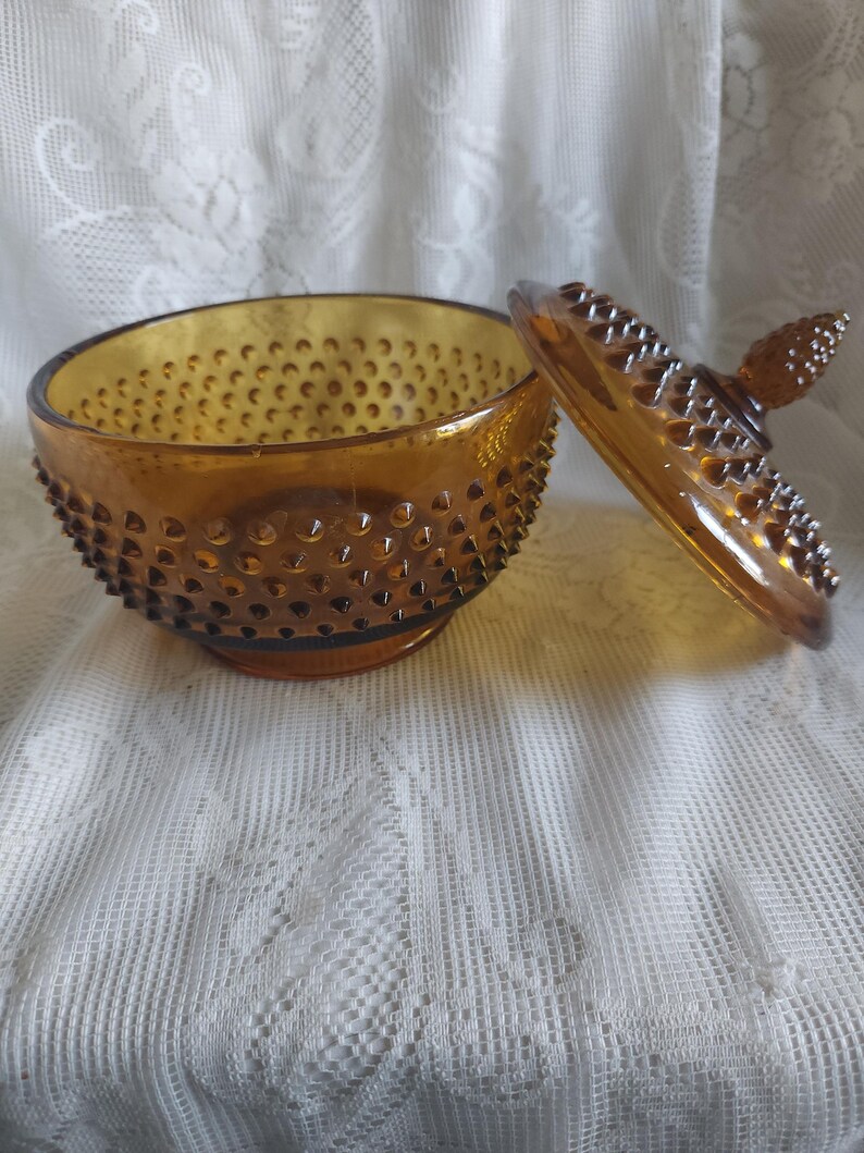 May include: Amber-colored glass candy dish with a hobnail pattern. The lid is off, revealing the interior. The dish sits on a white lace tablecloth. The lid has a knob on top.