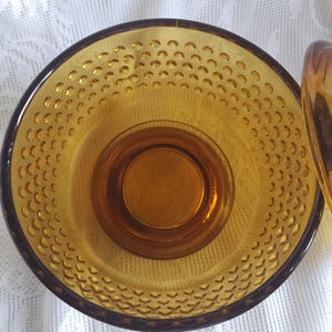 May include: Amber-colored glass bowl with a textured, dimpled interior. The bowl has a dark brown rim and a circular base. The design features a series of small, evenly spaced indentations across the surface.