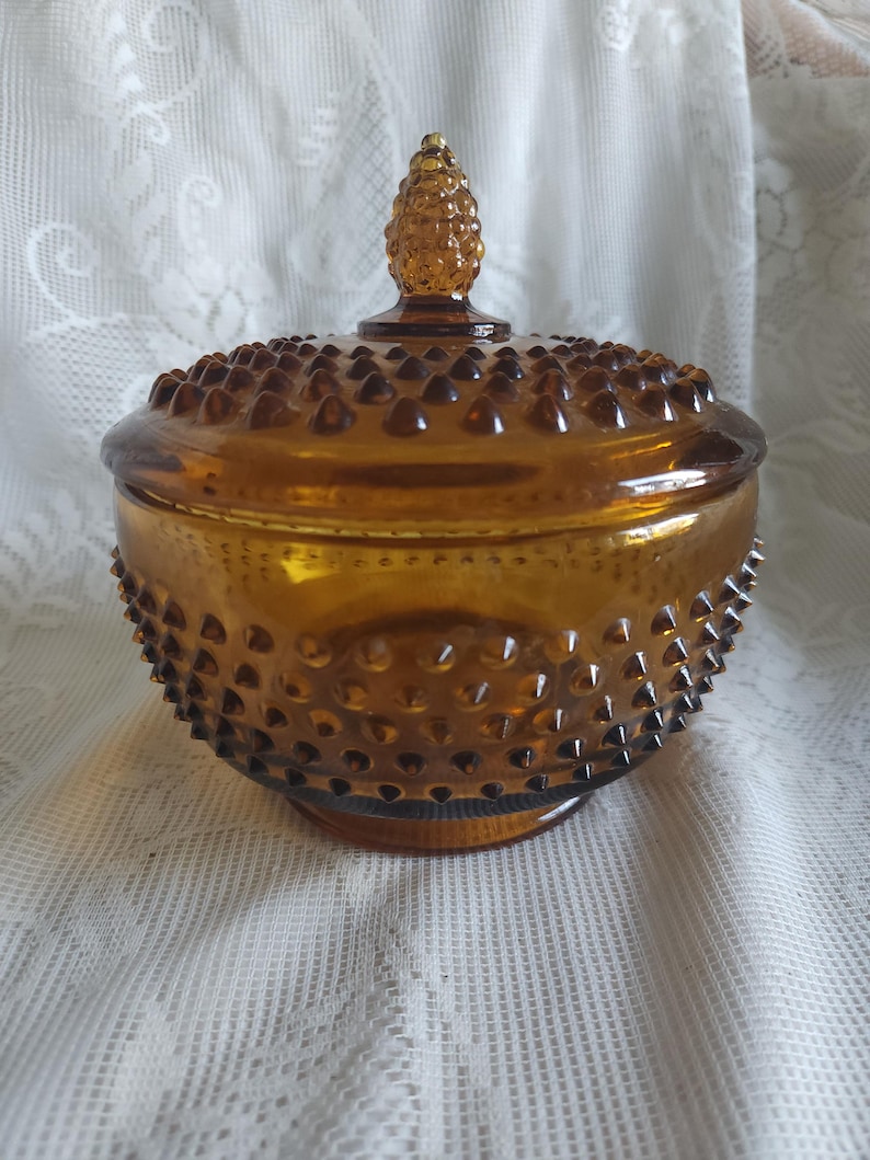 May include: Amber-colored glass candy dish with a lid. The dish has a hobnail pattern and a small, cone-shaped handle on the lid. The dish is round and sits on a small base. The glass has a warm, golden hue.