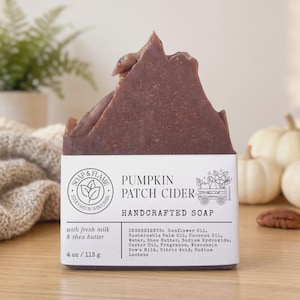 May include: A dark brown bar of handcrafted soap labeled Pumpkin Patch Cider. The white label features a circular logo, a wagon illustration, and text listing ingredients including sunflower oil, shea butter, and cow's milk, with a weight of 4 oz.