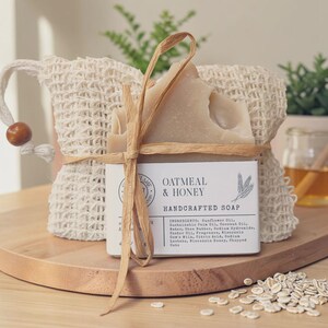 May include: A beige bar of oatmeal and honey handcrafted soap wrapped in a white paper label with black text and tied with a tan raffia ribbon, positioned in front of a textured, off-white mesh drawstring bag with a wooden bead detail.