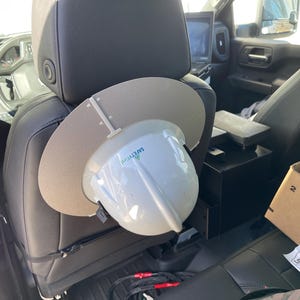 Może przedstawiać: Biały kask z szarym daszkiem i napisem "SAFETY FIRST" w zielonych literach jest przymocowany do zagłówka czarnego skórzanego fotela samochodowego.