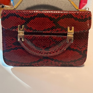 May include: A red and black snakeskin-patterned handbag with a rectangular shape. The bag features a top handle with gold-colored metal accents and a flap closure secured by two gold-colored clasps. The bag's design is classic and elegant.