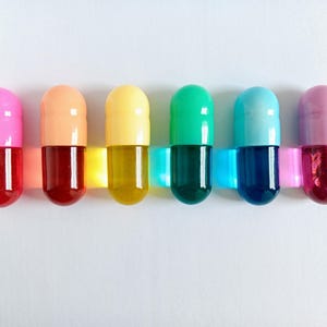 以下が含まれることがあります： 6つのカラフルなカプセル型のオブジェクト。それぞれ半透明の下半分と不透明な上半分があります。色はピンク、オレンジ、黄色、緑、青、紫です。透明なプラスチックストリップで接続されています。