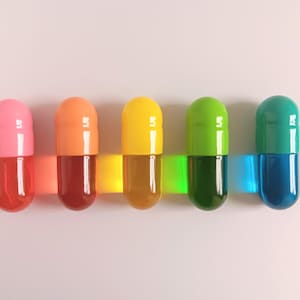May include: Five colorful capsule-shaped objects in pink, orange, yellow, green, and teal. Each capsule has a clear, liquid-filled lower half. The capsules are arranged horizontally, with a white background.