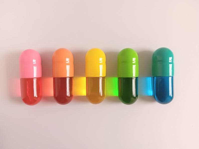 May include: Five colorful capsule-shaped objects in pink, orange, yellow, green, and teal. Each capsule has a clear, liquid-filled lower half. The capsules are arranged horizontally, with a white background.