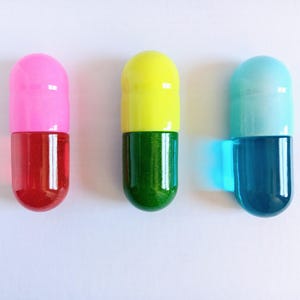 May include: Three colorful capsule-shaped objects. The first is pink and red, the second is yellow and green, and the third is light blue and dark blue. The objects are on a white background.