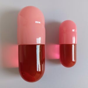 以下が含まれることがあります： 2つの大きく光沢のあるカプセル型の物体。上半分は薄いピンク色で、下半分は濃い赤茶色です。片方はもう片方よりかなり大きく、どちらも白い背景に対して垂直に配置されています。