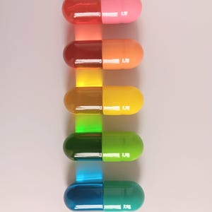 May include: A vertical arrangement of six colorful capsules, each split into two halves. The capsules transition from red and pink to orange and yellow, then to green and blue, creating a gradient effect. Each capsule has a small, white rectangular detail.