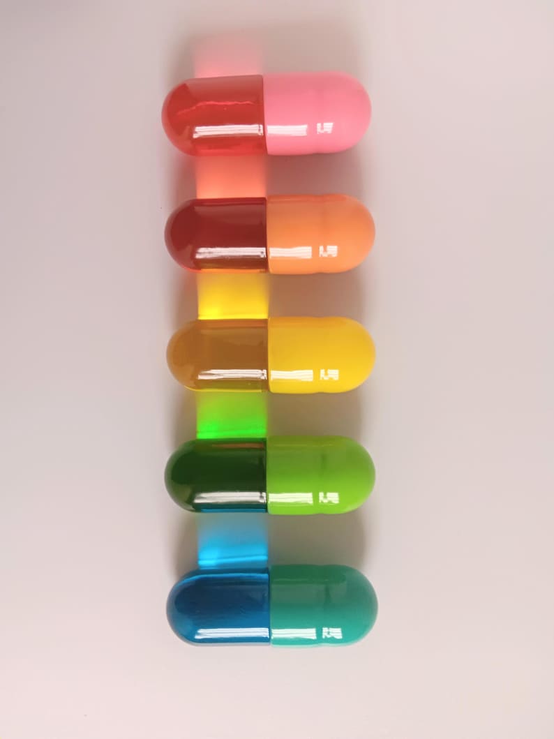 May include: A vertical arrangement of six colorful capsules, each split into two halves. The capsules transition from red and pink to orange and yellow, then to green and blue, creating a gradient effect. Each capsule has a small, white rectangular detail.