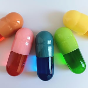 以下が含まれることがあります： オレンジ、ピンク、ティール、グリーン、イエローの色合いの、5つのカラフルな、特大のカプセル型のオブジェクト。各カプセルは透明で丸みを帯びた端と、単色のキャップが付いています。白い表面に配置されています。