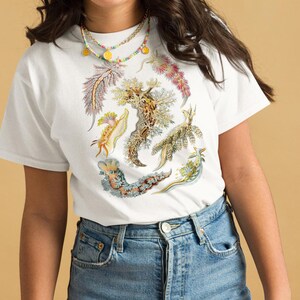 以下が含まれることがあります： カラフルな海のナメクジのグラフィックプリントが施された白いTシャツ。グラフィックには、黄色、オレンジ、ピンク、ブルーなど、さまざまな色の海のナメクジがさまざまなパターンで描かれています。海のナメクジは、シャツに円形に配置されています。