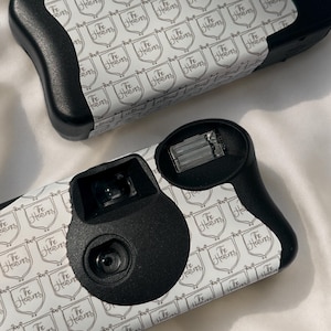 May include: Two black and white disposable cameras. The cameras have a repeating pattern of shields with the text "The Heavens". The cameras have black grips and lenses.